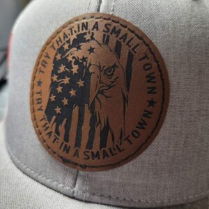Op de afbeelding: Een bruin leren patch met een Amerikaanse vlag en een adelaarontwerp. De patch heeft de tekst "Try that in a small town" rond de rand geschreven.