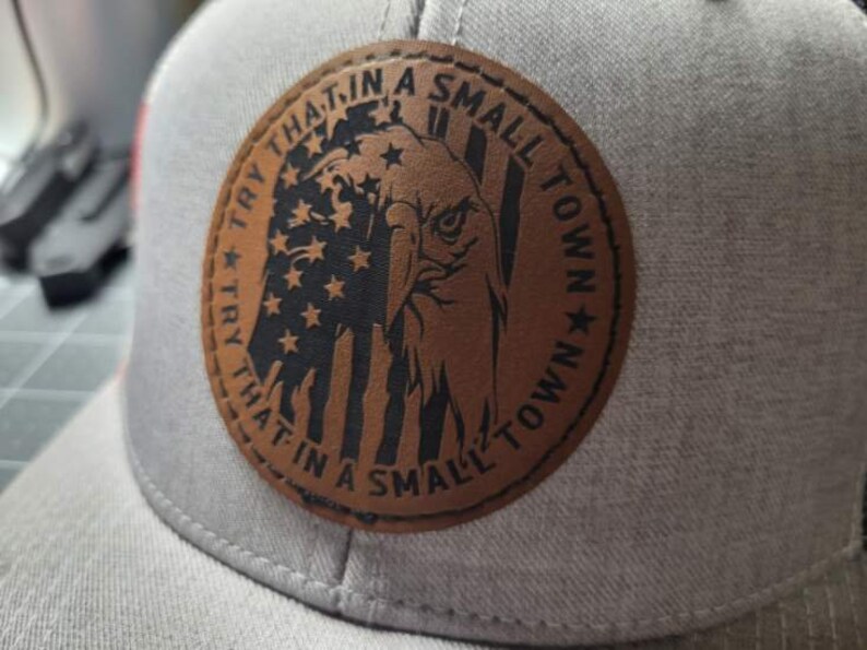 Op de afbeelding: Een bruin leren patch met een Amerikaanse vlag en een adelaarontwerp. De patch heeft de tekst "Try that in a small town" rond de rand geschreven.