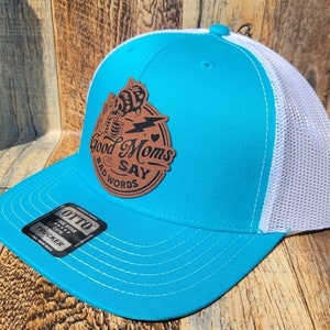 Peut inclure: Un chapeau de camionneur turquoise avec un filet blanc à l'arrière. Le chapeau présente un patch en cuir brun avec une abeille et le texte "Good Moms Say Bad Words".