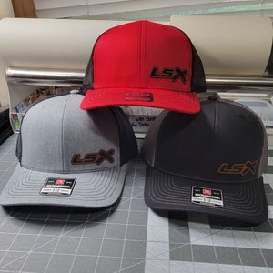 Peut inclure: Trois casquettes de baseball avec des panneaux arrière en maille noire. Les casquettes sont grises, noires et rouges. Chaque casquette a un patch en cuir marron avec le texte "LSX" sur le devant.
