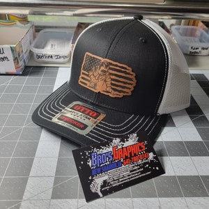 Può includere: Un cappello da camionista nero e bianco con una toppa in pelle marrone che presenta un saldatore con un design a bandiera americana. Il cappello ha una parte posteriore in rete bianca e le parole "OTTO FIVE STAR TRUCKER" sono stampate sul davanti.