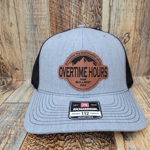 Puede incluir: Una gorra de camionero gris y negra con un parche de cuero marrón que dice "Been Sellin' My Soul Workin' all day" y "OVERTIME HOURS FOR BULLSHIT PAY". La gorra es ajustable y tiene una parte trasera de malla negra.