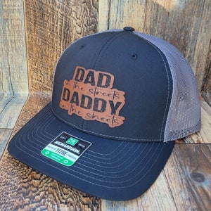 May include: A black and gray trucker hat with a brown leather patch that says "Dad in the streets Daddy in the sheets".