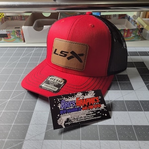 Peut inclure: Casquette de camionneur rouge et noire avec un patch en cuir marron sur lequel est inscrit "LSX". La casquette a un dos en maille noire et un devant rouge avec une visière noire cousue. La casquette a également une petite étiquette noire et blanche qui dit "Otto" et "Trucker".