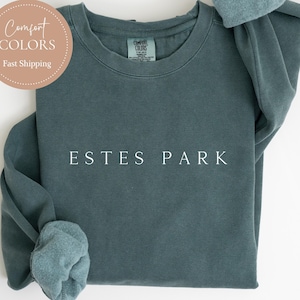 May include: A blue spruce crewneck sweatshirt with the text "ESTES PARK" in white. The sweatshirt has rolled-up sleeves and a round Comfort Colors logo. The words "BLUE SPRUCE" are at the bottom.