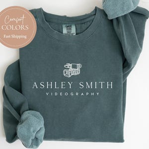 May include: Blue spruce crewneck sweatshirt with the text "ASHLEY SMITH VIDEOGRAPHY" and a camera graphic. The sweatshirt has rolled-up sleeves and a round Comfort Colors logo. The words "BLUE SPRUCE" are at the bottom.