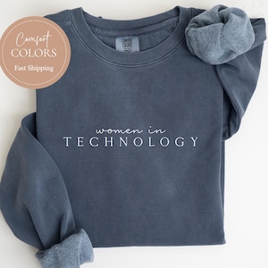 May include: A denim blue sweatshirt with the words "women in TECHNOLOGY" printed in white. The sweatshirt has a crew neck and long sleeves with cuffed wrists. A scrunchie rests on the shoulder. The words "Comfort Colors" and "Fast Shipping" are in a circle.