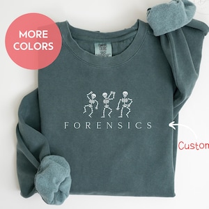 May include: Blue spruce sweatshirt with white skeleton graphic and the word "FORENSICS". The sweatshirt has rolled-up sleeves and a "MORE COLORS" button. The text "BLUE SPRUCE" is at the bottom.