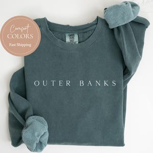 May include: A blue spruce sweatshirt with the text "OUTER BANKS" in white. The sweatshirt has a crew neck and long sleeves with folded cuffs. A circular logo in the upper left corner reads "Comfort Colors" and "Fast Shipping".