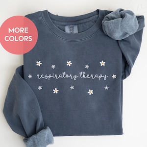 May include: A dark blue unisex sweatshirt with the text "respiratory therapy" in white cursive lettering and small white daisy flowers. The sweatshirt is made of a soft, comfortable material and is perfect for everyday wear.