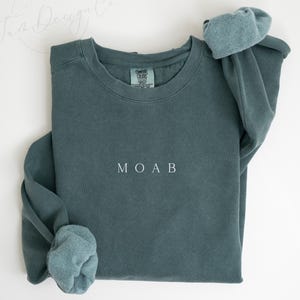 May include: A blue spruce sweatshirt with the word "MOAB" embroidered in white. The sweatshirt has a crew neck and long sleeves with cuffed wrists. The text "BLUE SPRUCE" is at the bottom of the image.