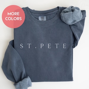 May include: A denim blue sweatshirt with the text "ST. PETE" in white. The sweatshirt has a crew neck and cuffed sleeves. A red circle with the words "MORE COLORS" is in the upper left corner. The word "DENIM" is in the lower right corner.