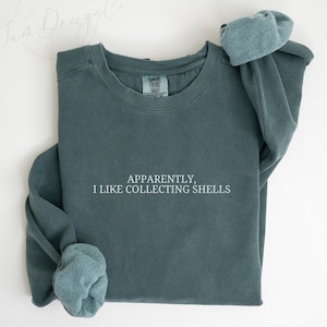 May include: Blue spruce sweatshirt with the text "Apparently, I like collecting shells" in white. The sweatshirt is folded with the sleeves rolled up. The words "Blue Spruce" are at the bottom of the image.