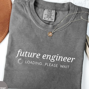 May include: A gray Comfort Colors t-shirt with the words "future engineer" and "LOADING...PLEASE WAIT" printed in white. A silver necklace with a small pendant rests on the shirt. The shirt is size S-P-CH.