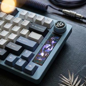 Könnte beinhalten: Hellblaue mechanische Tastatur mit weißen und blauen Tastenkappen. Die Tastatur hat eine dekorative Platte mit einem Koi-Fisch-Design und einen Lautstärkeregler. Ein Spiralkabel und eine dunkle Oberfläche sind ebenfalls sichtbar.