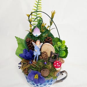 Op de afbeelding: Een sprookjesachtige tuin in een blauw-witte theekop. De miniatuurscène bevat een feeënbeeldje, blauwe bloemen, mos, dennenappels en een klein houten bankje. Een rode paddenstoel met witte stippen.