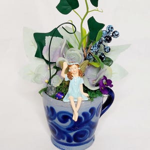 May include: A blue ceramic teacup with a decorative design, filled with greenery, flowers, and a fairy figurine. The fairy is wearing a light blue dress and sitting on the rim of the cup. The arrangement includes green leaves, purple flowers, and blue berries.