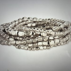May include: A multi-strand bracelet composed of numerous small, metallic silver-toned beads. The beads feature irregular, organic shapes with a polished, reflective finish, creating a layered, textured appearance across the entire circular piece.