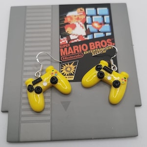 Op de afbeelding: Een paar gele videogamecontroller-oorbellen met zwarte knoppen en zilveren haken. De oorbellen worden getoond op een Nintendo Entertainment System-gamecartridge-doos met het Super Mario Bros.-logo.