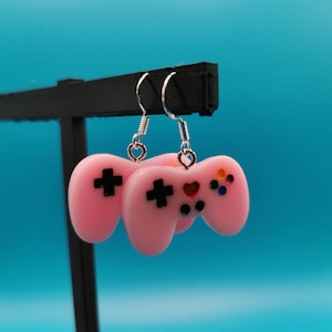 Op de afbeelding: Roze oorbellen in de vorm van een videogamecontroller met zwarte kruisknoppen en kleurrijke knoppen. De oorbellen hangen aan zilveren haken. De achtergrond is een blauwe gradiënt.