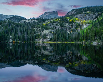 May include: A mountain lake with a reflection of the surrounding forest and a mountain peak in the water. The sky is a vibrant pink and blue.
