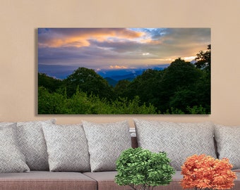 May include: A panoramic view of a mountain range at sunset, with a cloudy sky in shades of pink, orange, and blue. The mountains are covered in lush green trees, and the scene is framed by a few trees in the foreground.