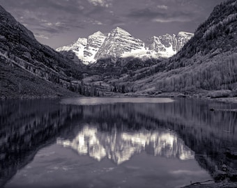 May include: Black and white landscape photograph of a mountain range reflected in a calm lake. The snow-capped peaks are centered, with forested slopes on either side. The sky is overcast, creating a dramatic, moody scene.