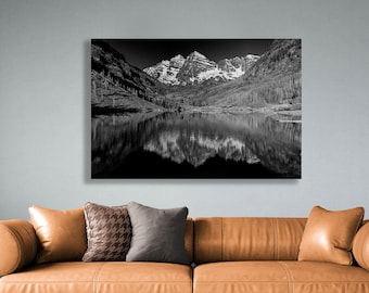 May include: Black and white landscape photograph of a mountain range reflected in a lake. The peaks are snow-capped, and the water mirrors the mountains and surrounding trees. The image is mounted on a wall above a brown leather couch.