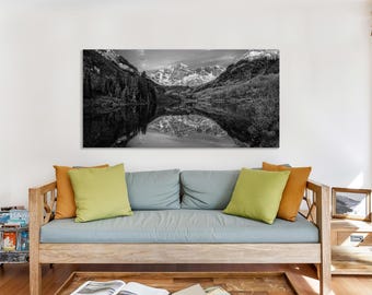 May include: Black and white landscape photograph of a mountain range reflected in a lake. The image is mounted above a wooden sofa with colorful pillows. The scene captures the natural beauty of the mountains and water.
