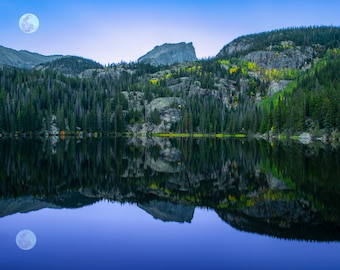 May include: A full moon hangs in the sky above a still lake reflecting the surrounding mountains and trees. The water is a deep blue, and the mountains are a dark green.