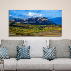 Pode incluir: Uma fotografia panorâmica de uma paisagem montanhosa com um céu azul e nuvens brancas. As montanhas são cobertas de árvores verdes e alguma neve. O primeiro plano é um campo gramado.