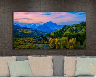 Könnte beinhalten: Ein Panoramafoto einer Bergkette mit einem Tal im Vordergrund. Die Berge sind blau und schneebedeckt, und das Tal ist mit Bäumen in Grün- und Gelbtönen gefüllt. Der Himmel ist in sanftem Rosa und Blau gehalten.