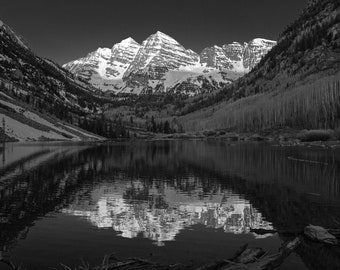 May include: A black and white photograph of a mountain range reflected in a still lake. The mountains are snow-capped and the lake is surrounded by trees.