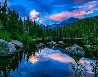 May include: A serene mountain lake reflecting a vibrant sunset sky. The water is still and clear, with a few large rocks scattered along the shore. The surrounding forest is lush and green, with tall pine trees lining the banks. The mountains in the background are majestic and imposing, with snow-capped peaks.