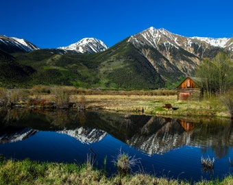 May include: A wooden cabin with a red door sits in a grassy field with a pond reflecting the snow-capped mountains in the background.