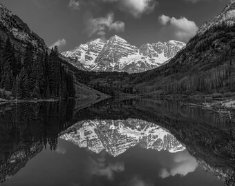 May include: Black and white landscape photograph of a mountain range reflected in a lake. The snow-capped peaks are mirrored in the still water, with dark forests lining the slopes. The sky is partially cloudy.