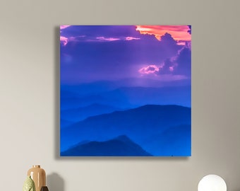 May include: A photograph of a mountain range at sunset. The sky is a vibrant purple and pink, with clouds casting long shadows over the blue mountains.