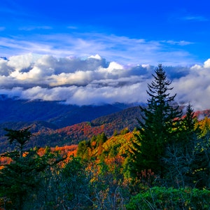 May include: A scenic view of a mountain range with fall foliage in shades of red, orange, and yellow. The mountains are covered in clouds, and the sky is a bright blue.