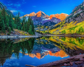 Puede incluir: Una vista panorámica de un lago de montaña con el reflejo de las montañas y los árboles circundantes. El agua es de un azul profundo, y las montañas son una mezcla de verde y rojo. Los árboles son una mezcla de verde y amarillo, y el cielo es de un azul brillante.