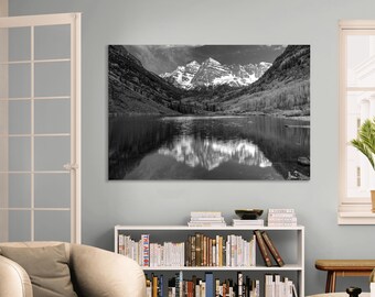 May include: Black and white landscape photograph of a mountain range reflected in a calm lake. The snow-capped peaks are framed by dark cliffs and trees, creating a dramatic and serene scene. The image is mounted on a wall.