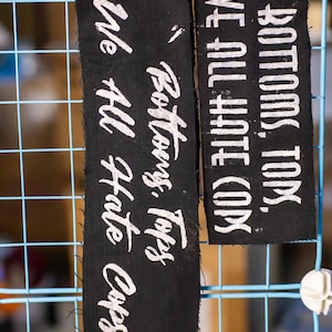 May include: Two black fabric patches with white text. One reads "BOTTOMS, TOPS, WE ALL HATE COPS." The other says "Bottoms, Tops, We All Hate Cops." Both are hanging on a blue wire grid.