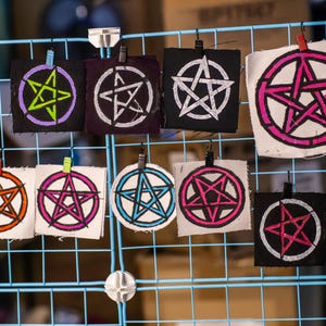 May include: A collection of square fabric patches, each featuring a pentagram design in various colors, including purple, orange, pink, and blue. The patches are displayed on a blue wire grid, held by small clips.
