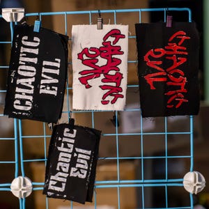 May include: Four fabric patches with text are displayed on a blue grid. Two black patches read "CHAOTIC EVIL" in white. One white patch and one black patch read "CHAOTIC" in red. The patches are held by small clips.