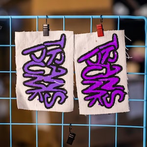 May include: Two rectangular patches with the word "PUNKS" in purple, outlined in black, are displayed. The patches are attached to a light blue grid with black and red clips. The background is blurred.