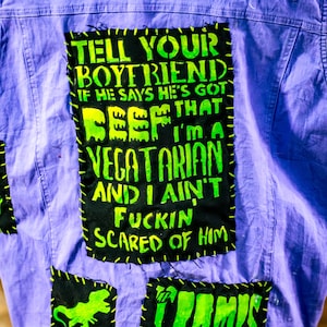 May include: A purple denim jacket with patches. The largest patch has the text "TELL YOUR BOYFRIEND IF HE SAYS HE'S GOT BEEF THAT I'M A VEGETARIAN AND I AIN'T FUCKIN SCARED OF HIM" in green and yellow. Other patches include a green dinosaur and the word "CRAMPS".