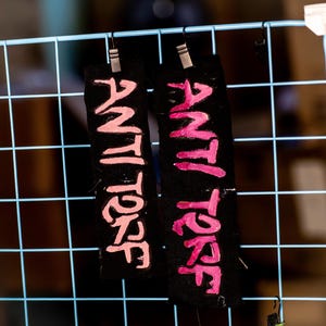 May include: Two black fabric tags with the words "ANTI TORF" in pink, handwritten-style lettering. The tags are hanging from a blue wire grid with small metal clips. The background is blurred.