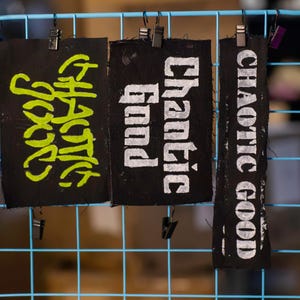 May include: Three black fabric patches with text are clipped to a blue wire grid. The left patch has the words "CHAOTIC GOOD" in neon green. The middle patch reads "Chaotic Good" in white. The right patch says "CHAOTIC GOOD" in white.