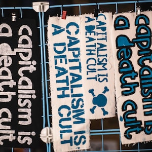 May include: Three fabric banners with the text "capitalism is a death cult" in white and blue lettering. The banners are hanging on a blue grid. One banner is black, and the other two are white.