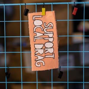 May include: A peach-colored fabric tag with the words "SUPPORT LOCAL DRAG" in bold, white capital letters. The tag is attached to a blue wire grid with small, colorful clips. The background is blurred, suggesting an indoor setting.