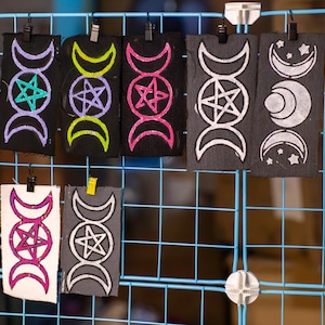 May include: A collection of rectangular fabric pieces with celestial designs, including crescent moons and stars, and pentagrams. The designs are in various colors, such as purple, green, pink, and white, against black and gray backgrounds. The pieces are clipped to a blue grid.